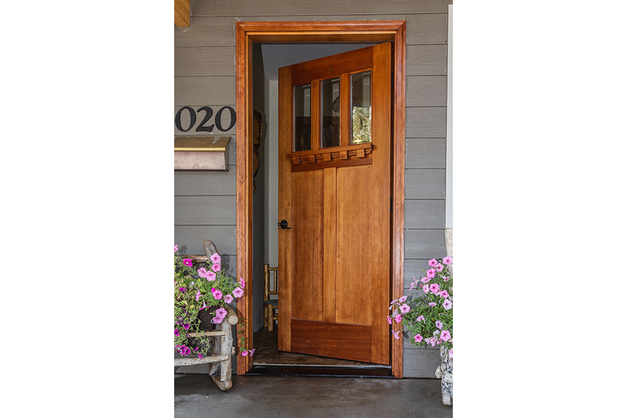 A brown wooden door sits ajar, revealing a minimal threshold. On each side of the door, on the porch, sit pots full of small pink flowers.